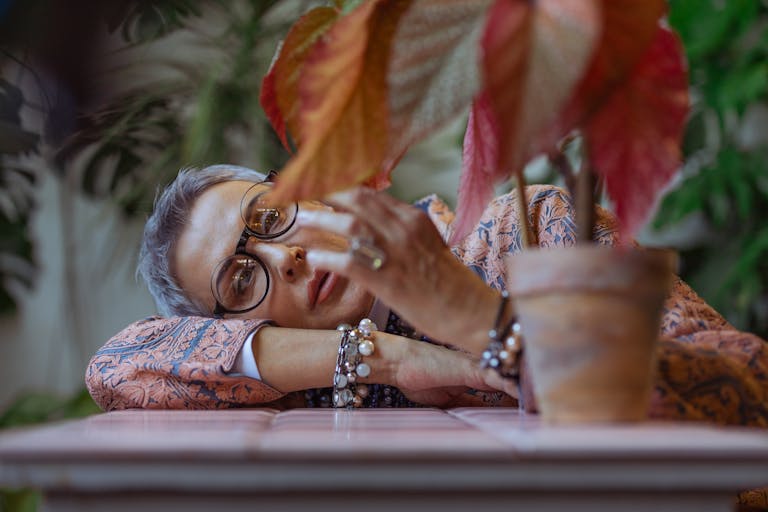 Elderly woman with eyeglasses and bracelets, pensive in a boho style setting with a decorative plant.