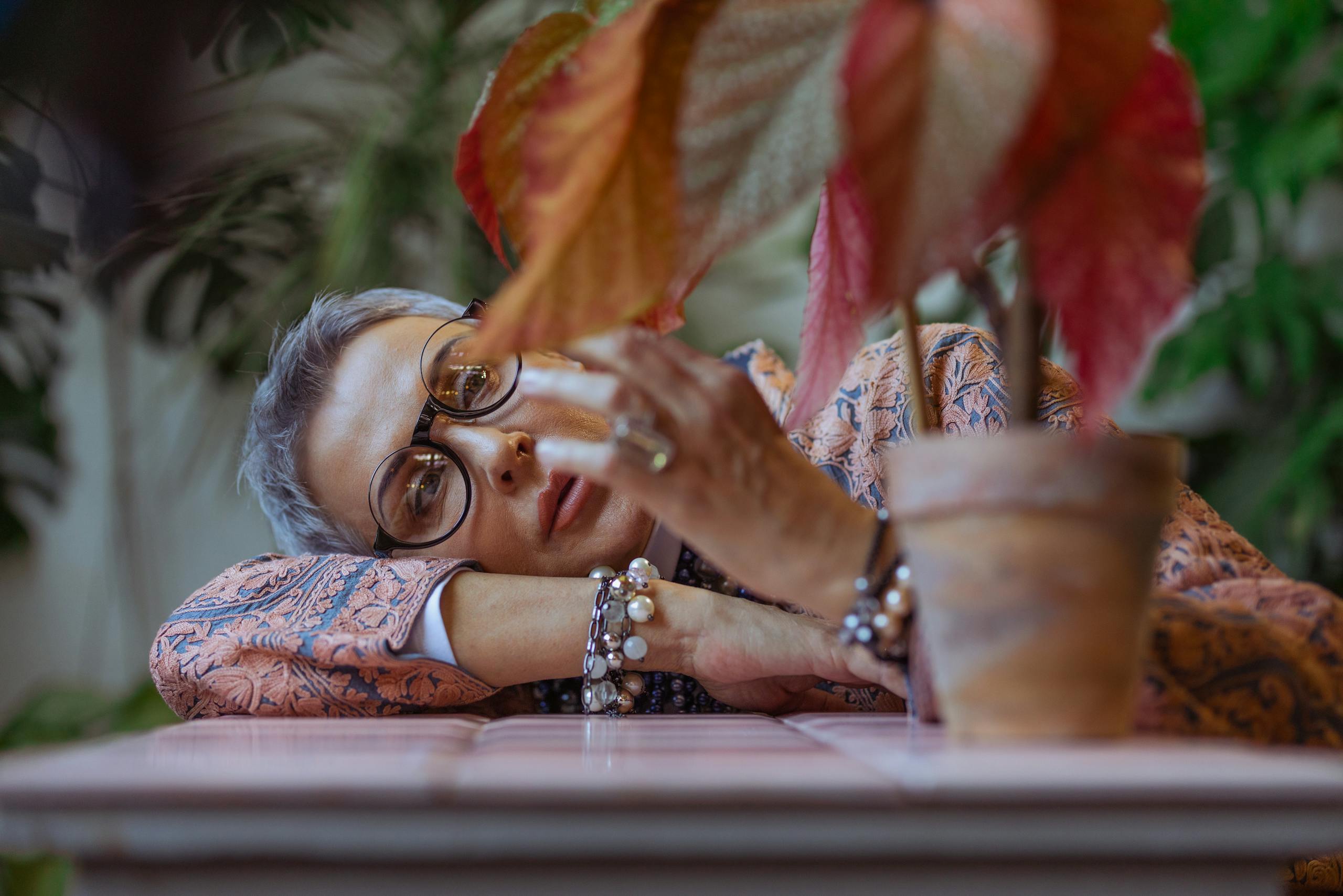 Elderly woman with eyeglasses and bracelets, pensive in a boho style setting with a decorative plant.